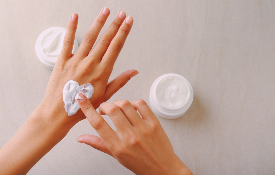 Der Zeigefinger einer rechten Hand formt auf dem Handrücken der linken Hand ein Herz aus weißer Creme. Im Hintergrund sind zwei weiße Cremetiegel zu sehen.