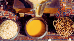 Auf einem orientalischen Teppich steht eine traditionelle Ölmühle, aus der goldgelbes Öl in eine Schale fließt. Links und rechts davon steht jeweils eine Schale mit vorbereiteten und rohen Argannüssen.  