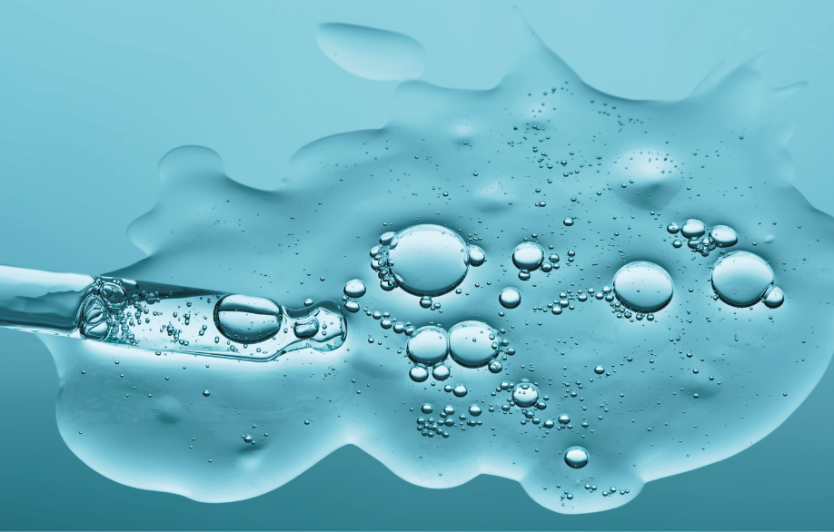 Die Detailaufnahme einer gläsernen Pipette, die eine durchsichtige Flüssigkeit mit Bläschen auf einen helltürkisblauen Hintergrund spritzt.